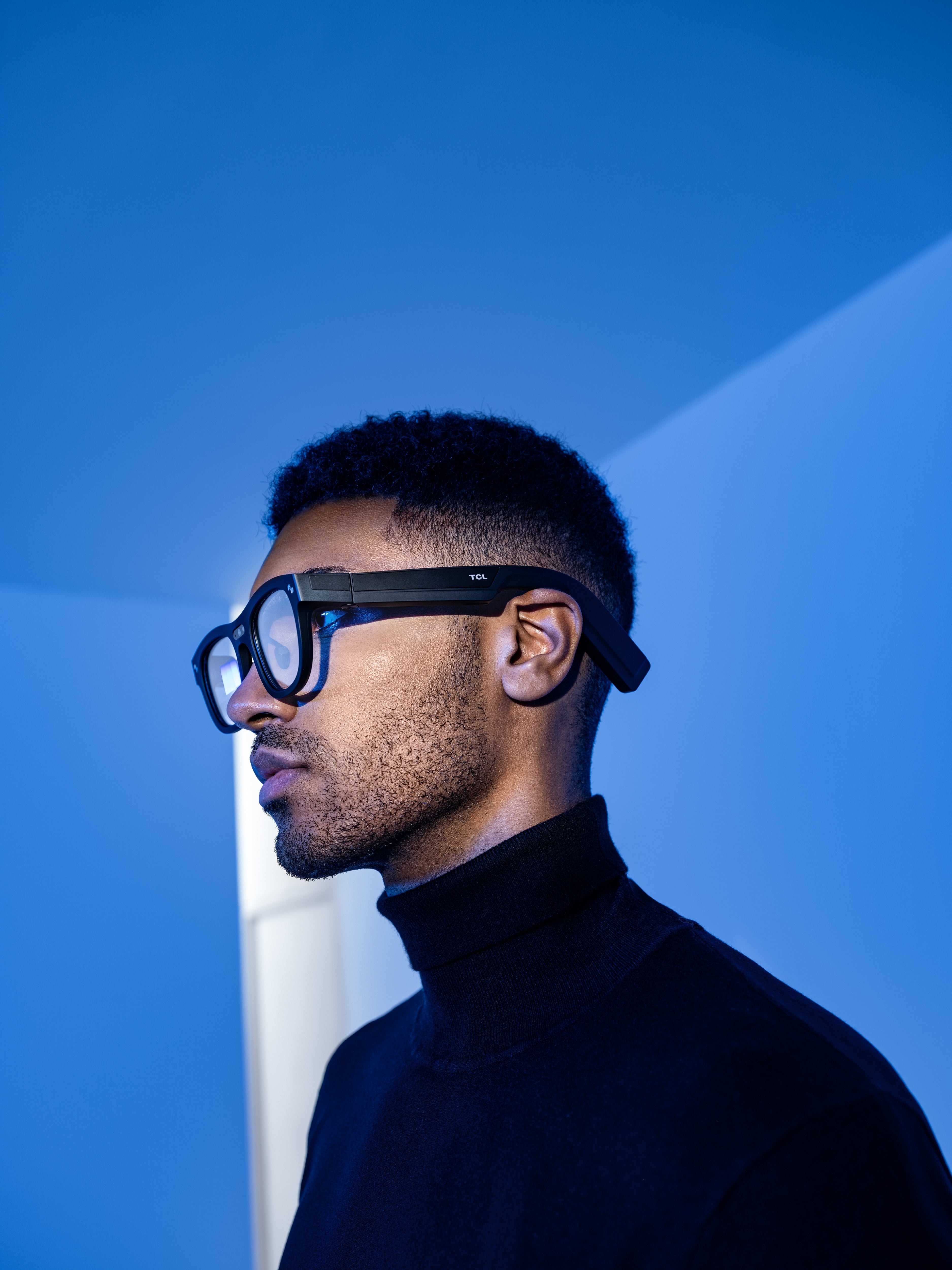 Man wearing black-framed TCL smart glasses and a black turtleneck in a blue room.